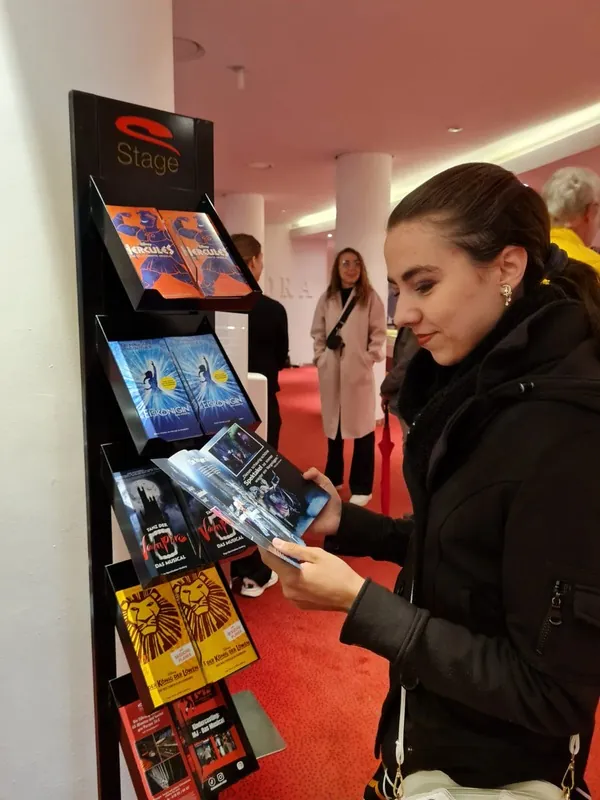 Vicky blättert an einem Ständer mit Programmheften im Theaterfoyer