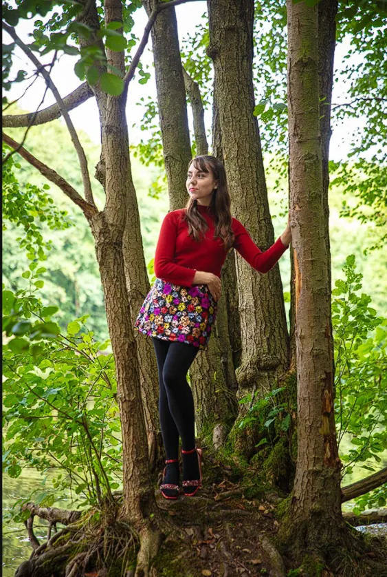 Vicky im roten Pullover und bunten Rock zwischen Baumstämmen